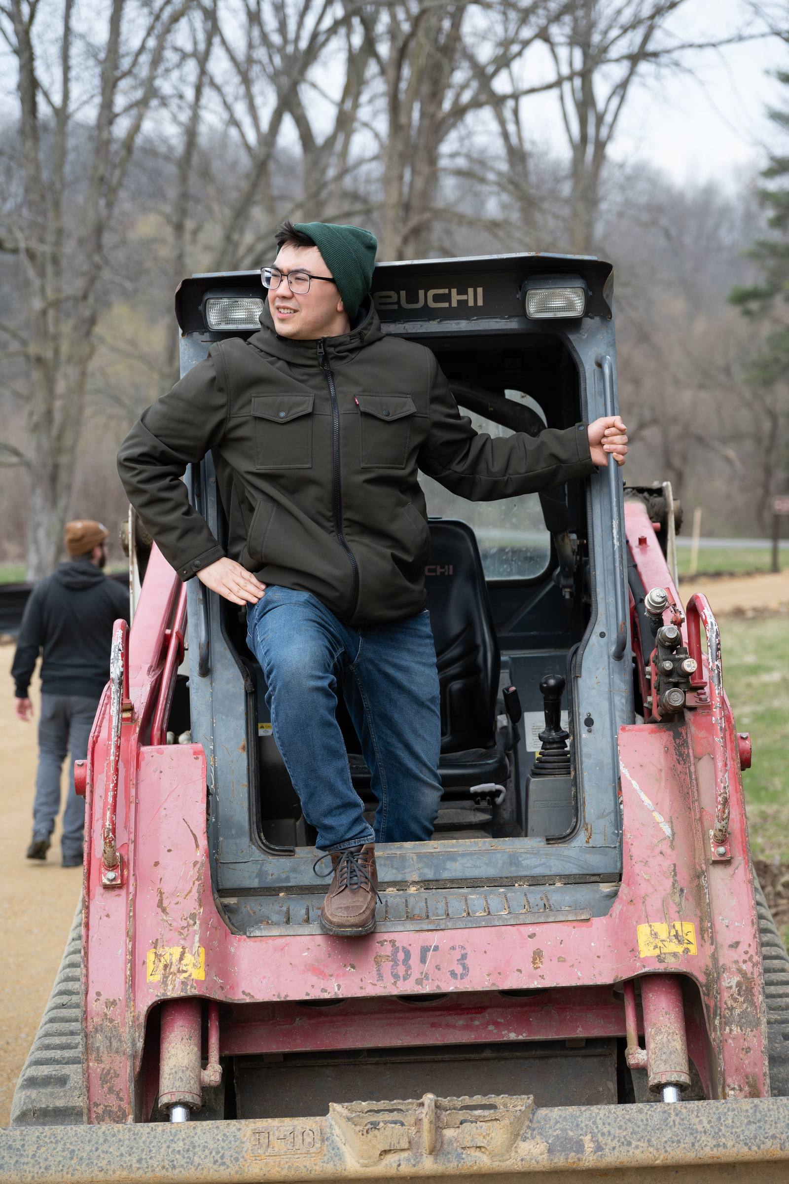 A person rides on the front of a tractor in a wooded work area.