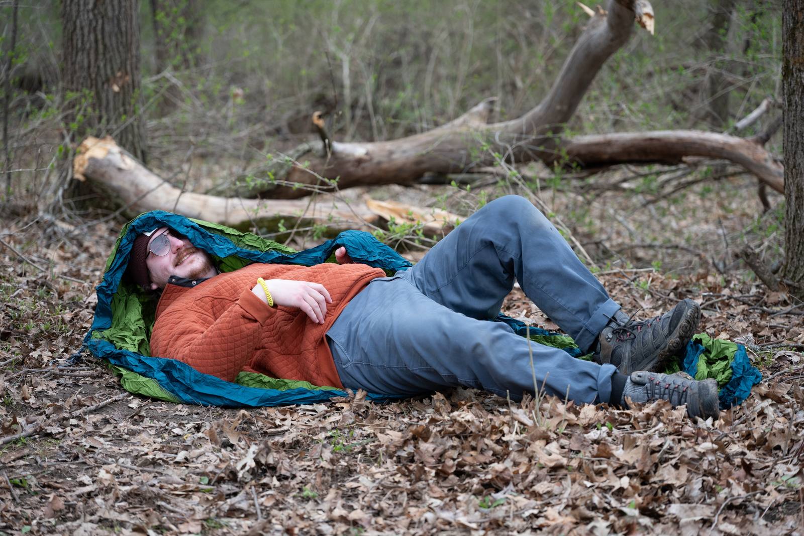 A man rests on a blanket in the woods beside fallen branches.