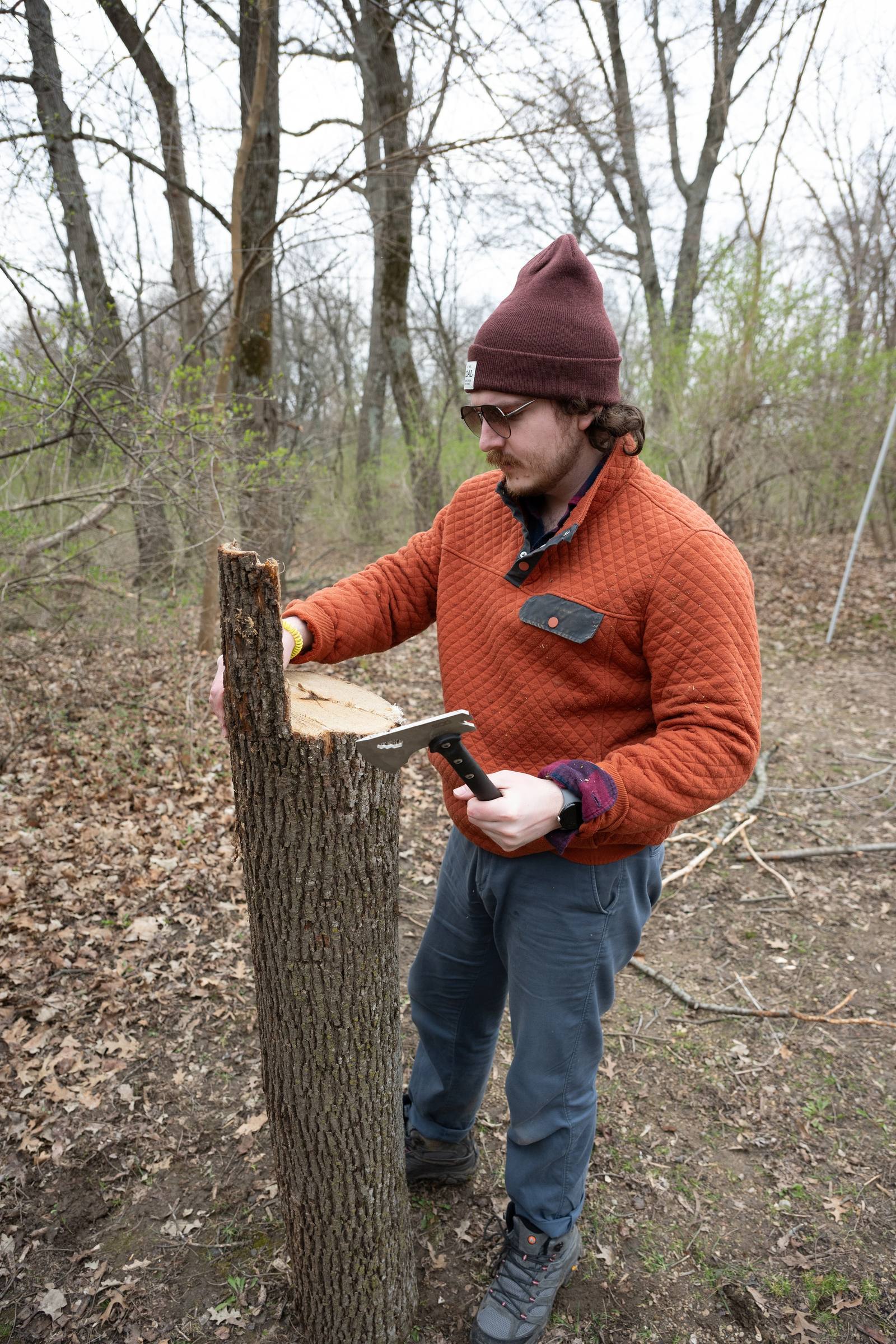 A person in an orange jacket works beside a tree in the woods.