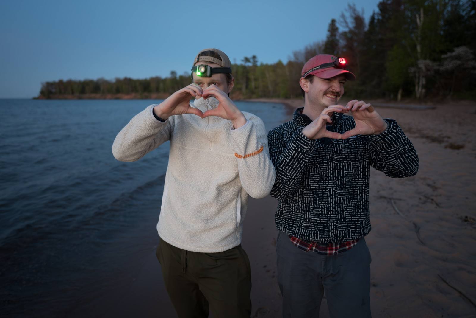 Two people wearing headlamps stand on a sandy lakeshore at dusk, each forming a heart shape with their hands.