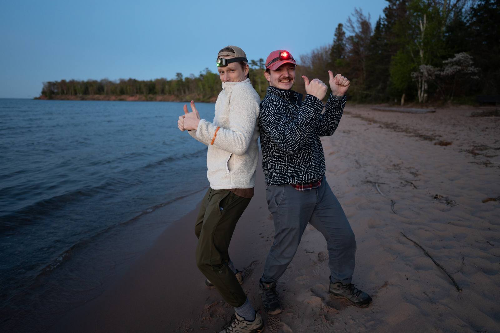 Two people in headlamps stand back-to-back on a sandy lakeshore at dusk, each giving a thumbs up toward the camera.