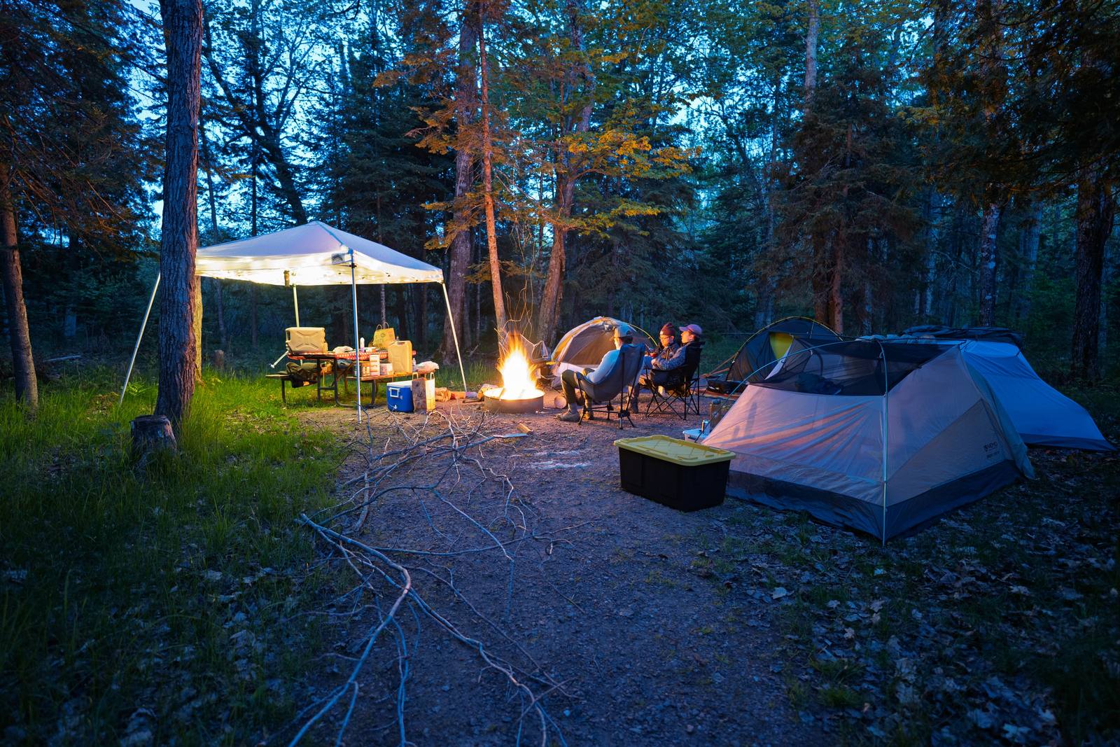 Three campers sit around a glowing campfire between two tents at dusk, beneath a lit canopy shelter in a wooded campsite.