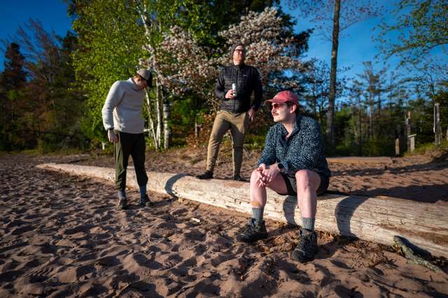 Three people gather on a sandy lakeshore beside a large driftwood log on a sunny spring day, framed by birch trees and a blooming white flowering tree.