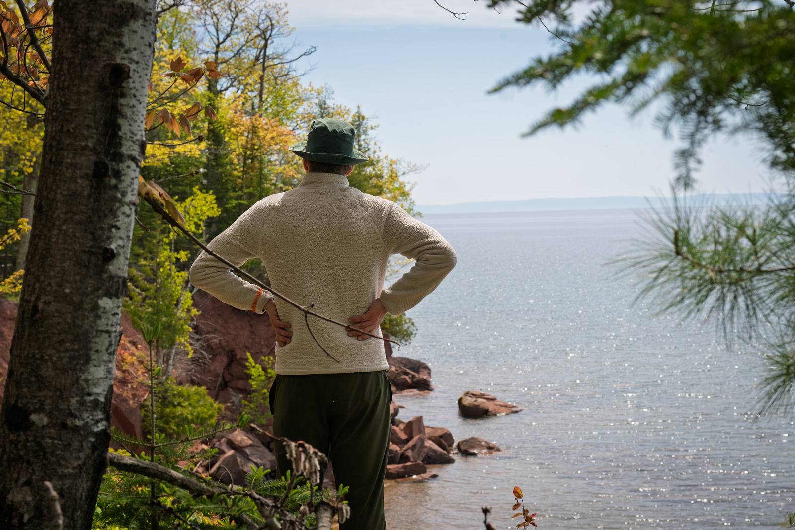 A person in a cream fleece and green bucket hat stands with hands on hips, looking out across a sparkling lake from a rocky red sandstone shore framed by birch trees and pines in early autumn.