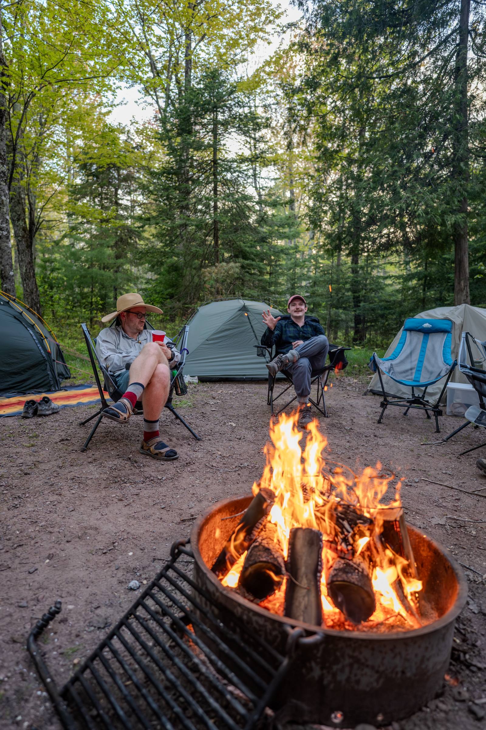 Two campers relax in chairs around a roaring campfire at a wooded campsite, surrounded by tents and tall pines on a spring evening.