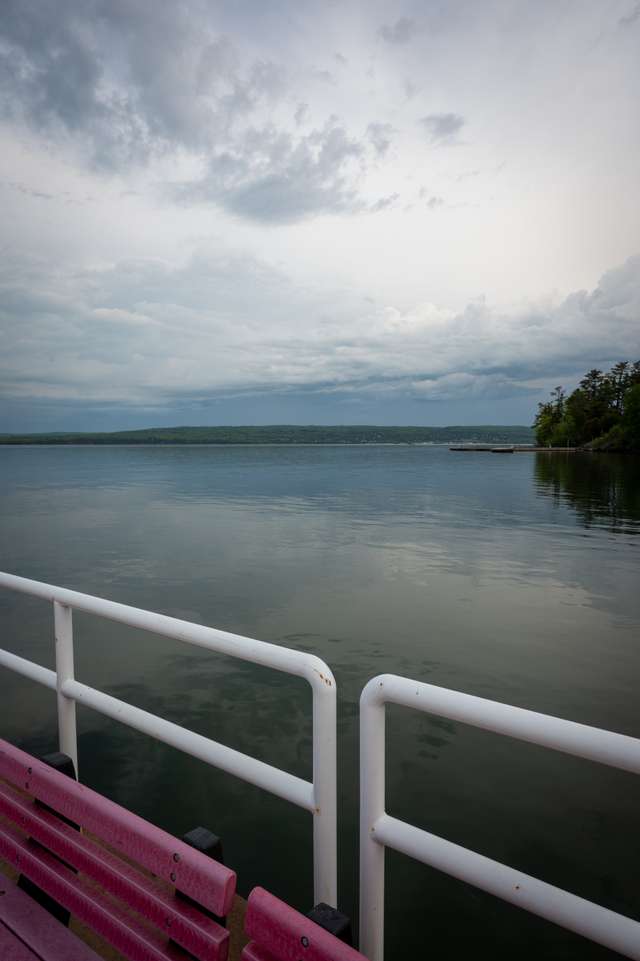 A pink bench on a dock with white railings looks out over a calm lake under a stormy sky, with a forested island on the right and green hills across the water.