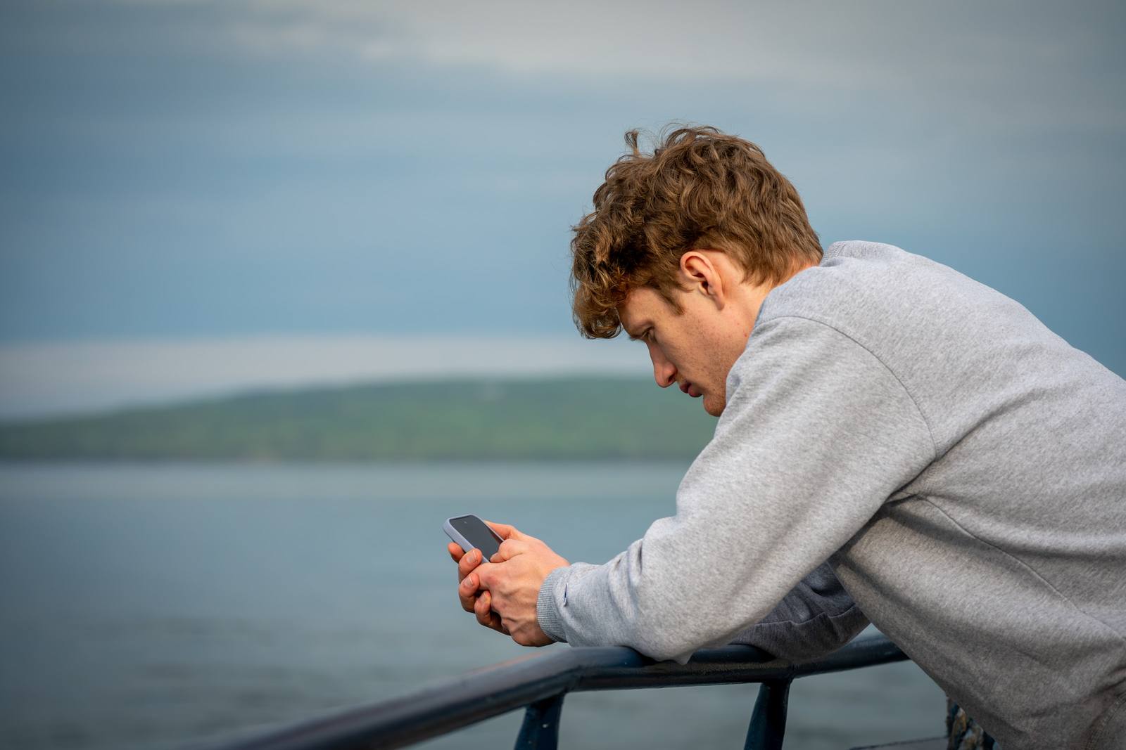 A young man in a grey sweatshirt leans on a ferry railing and looks at his phone, with a green-forested island visible across calm water in the background.