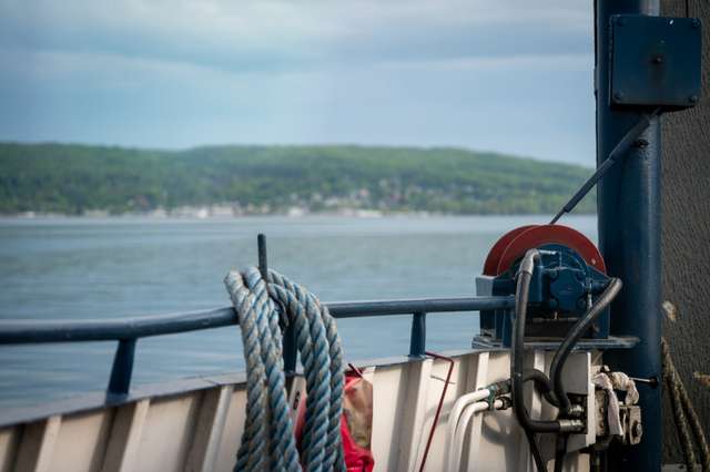 Heavy nautical rope and a deck winch on a ferry, with a green forested shoreline and small harbor visible across calm water in the distance.