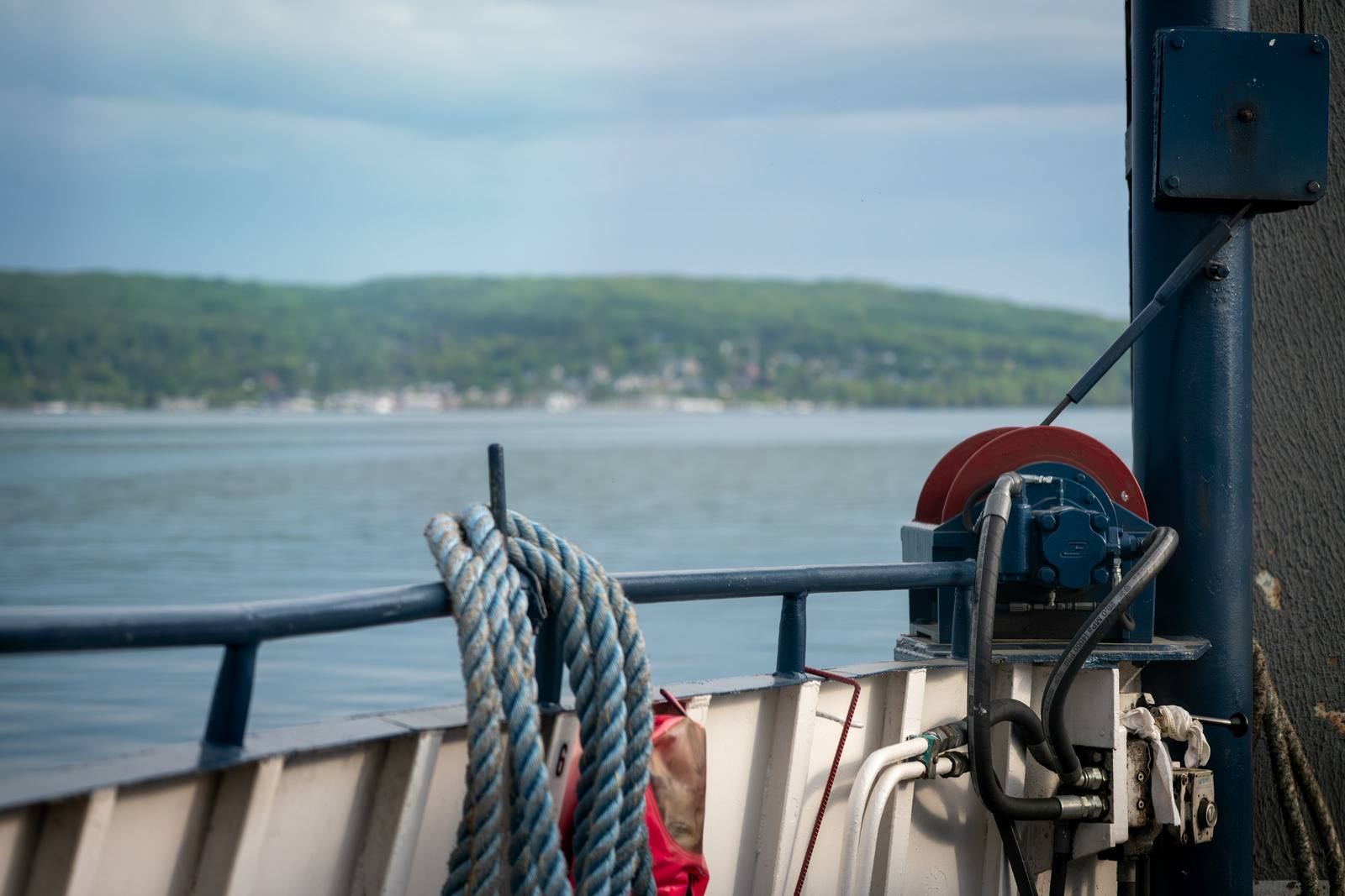 Heavy nautical rope and a deck winch on a ferry, with a green forested shoreline and small harbor visible across calm water in the distance.
