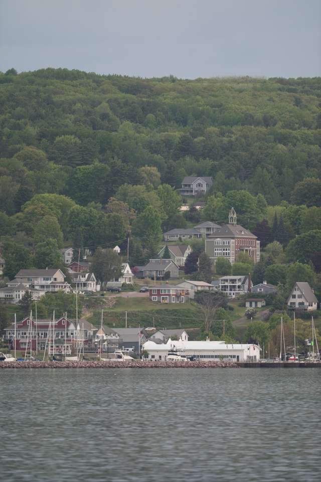 A lakeside town viewed from the water, with a marina full of sailboat masts, colorful storefronts, and a historic brick Victorian building set against a forested hillside.