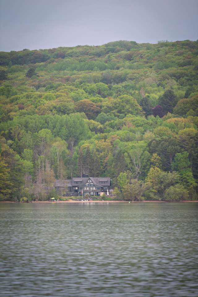 A dark craftsman-style lakehouse sits at the water's edge, nestled against a dense spring-green hillside viewed from across a calm lake.