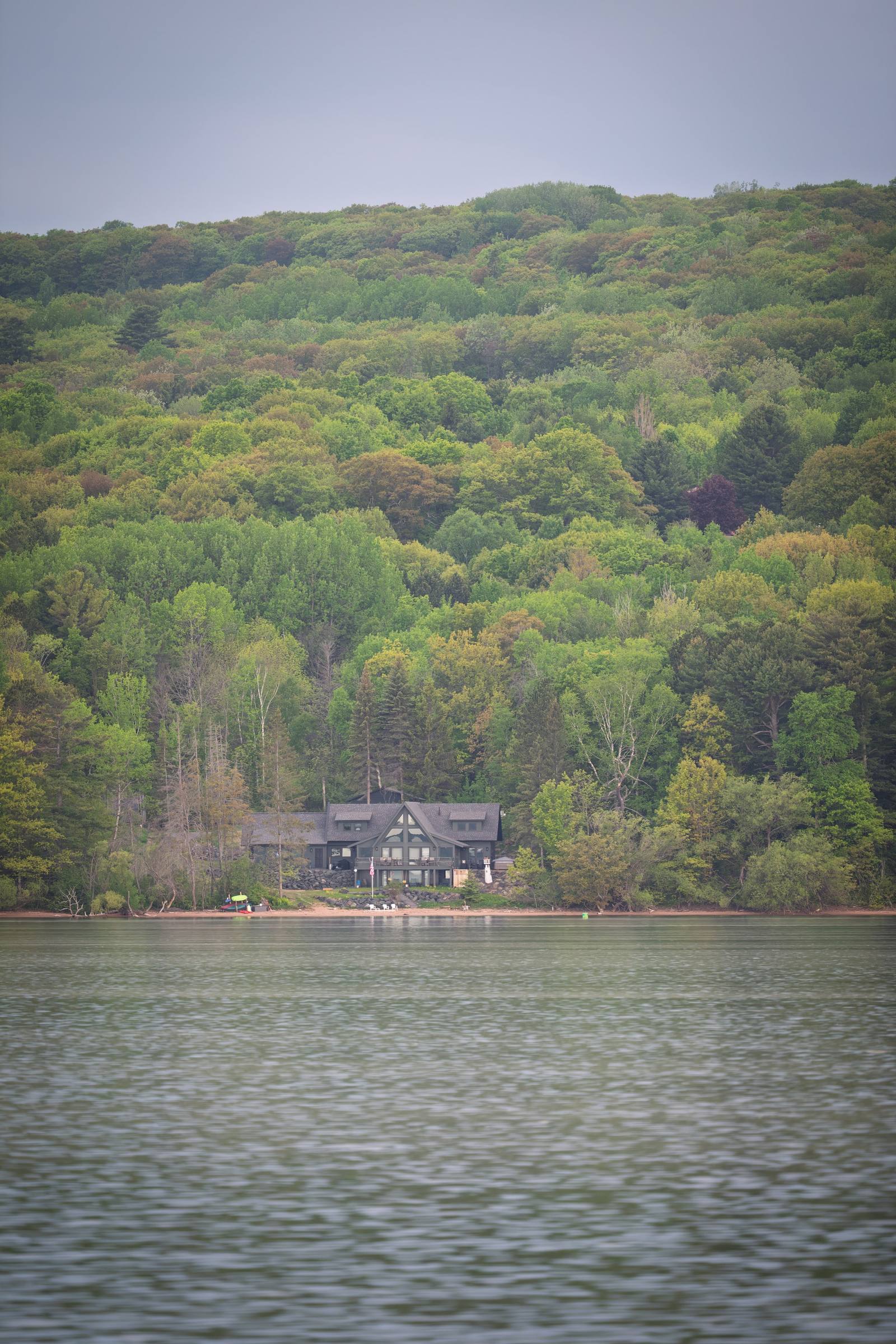 A dark craftsman-style lakehouse sits at the water's edge, nestled against a dense spring-green hillside viewed from across a calm lake.